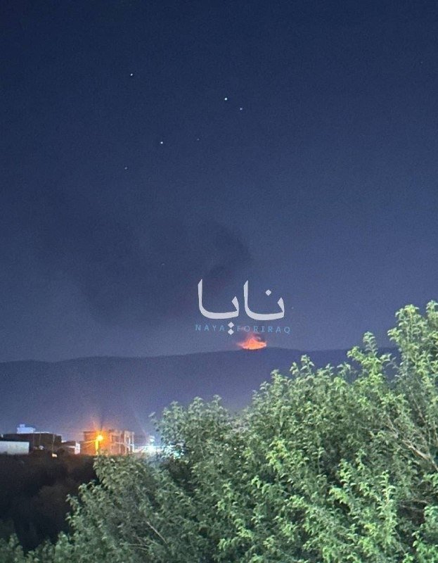 Nighttime view of a distant hillside fire with smoke rising, scattered lights, foreground trees, and overlaid Kurdish and English text reading "Naya For Iraq."