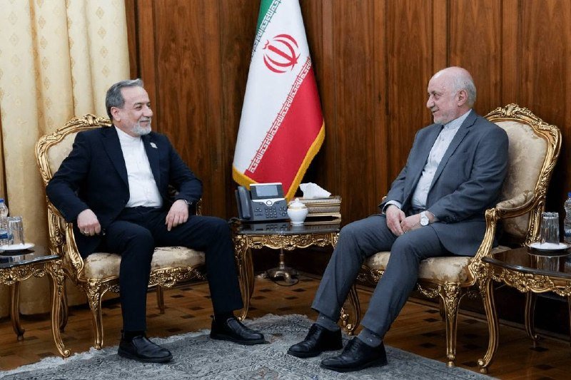 Two bearded men in suits sit in ornate chairs facing each other in a wood-paneled room, with the Iranian flag displayed between them.