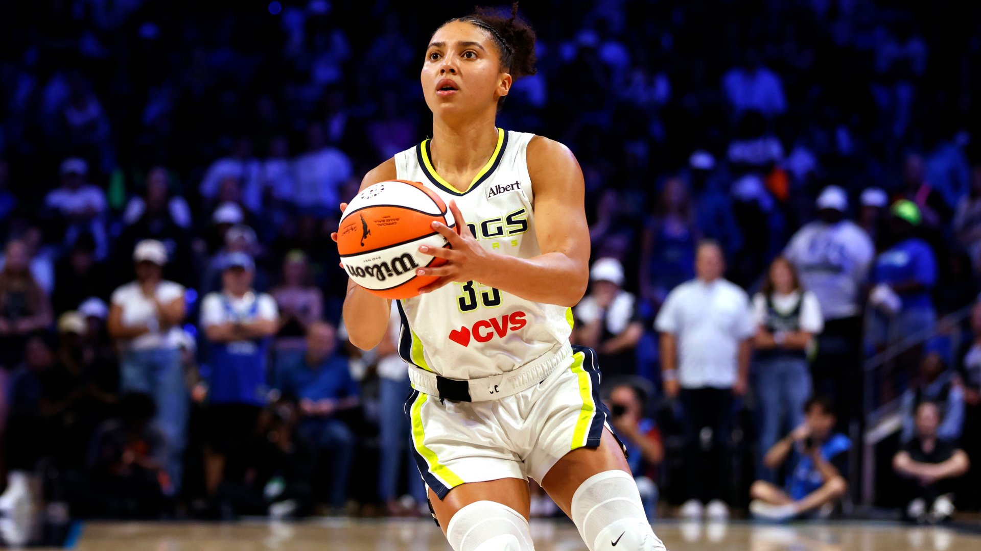 A basketball player in a white "Wings" jersey with CVS sponsorship prepares to shoot a basketball, with a blurred crowd in the arena background.