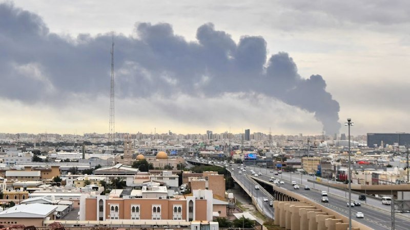 A multi-lane highway with vehicles cuts through a sprawling cityscape, while thick plumes of dark smoke rise into the cloudy sky on the horizon.