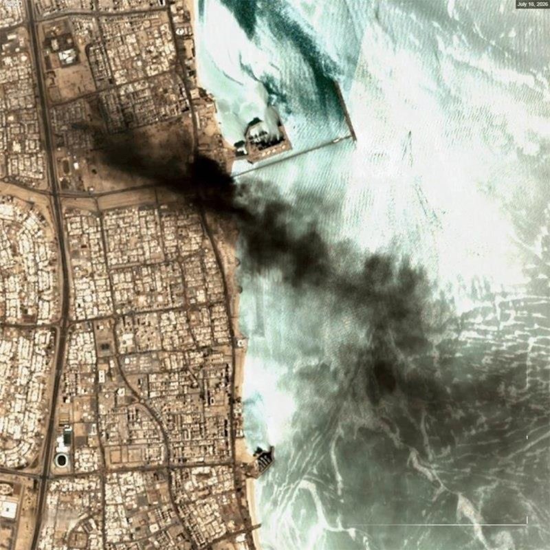 Satellite image dated July 18, 2020, shows dark smoke billowing across a coastline from two structures, one extending into the turquoise water, adjacent to a densely built urban grid.