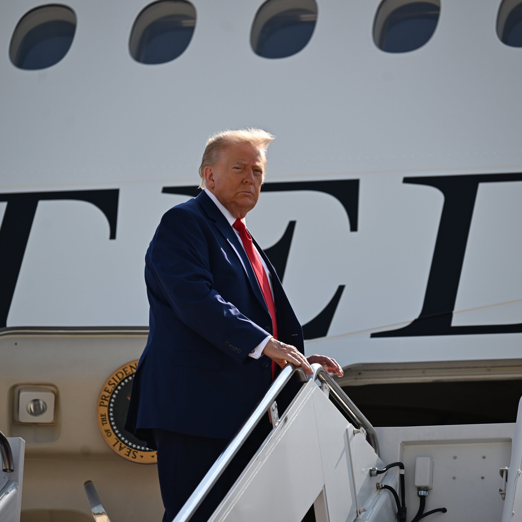 A man with blonde hair, wearing a dark blue suit and red tie, descends the staircase of a white aircraft displaying the Presidential Seal.