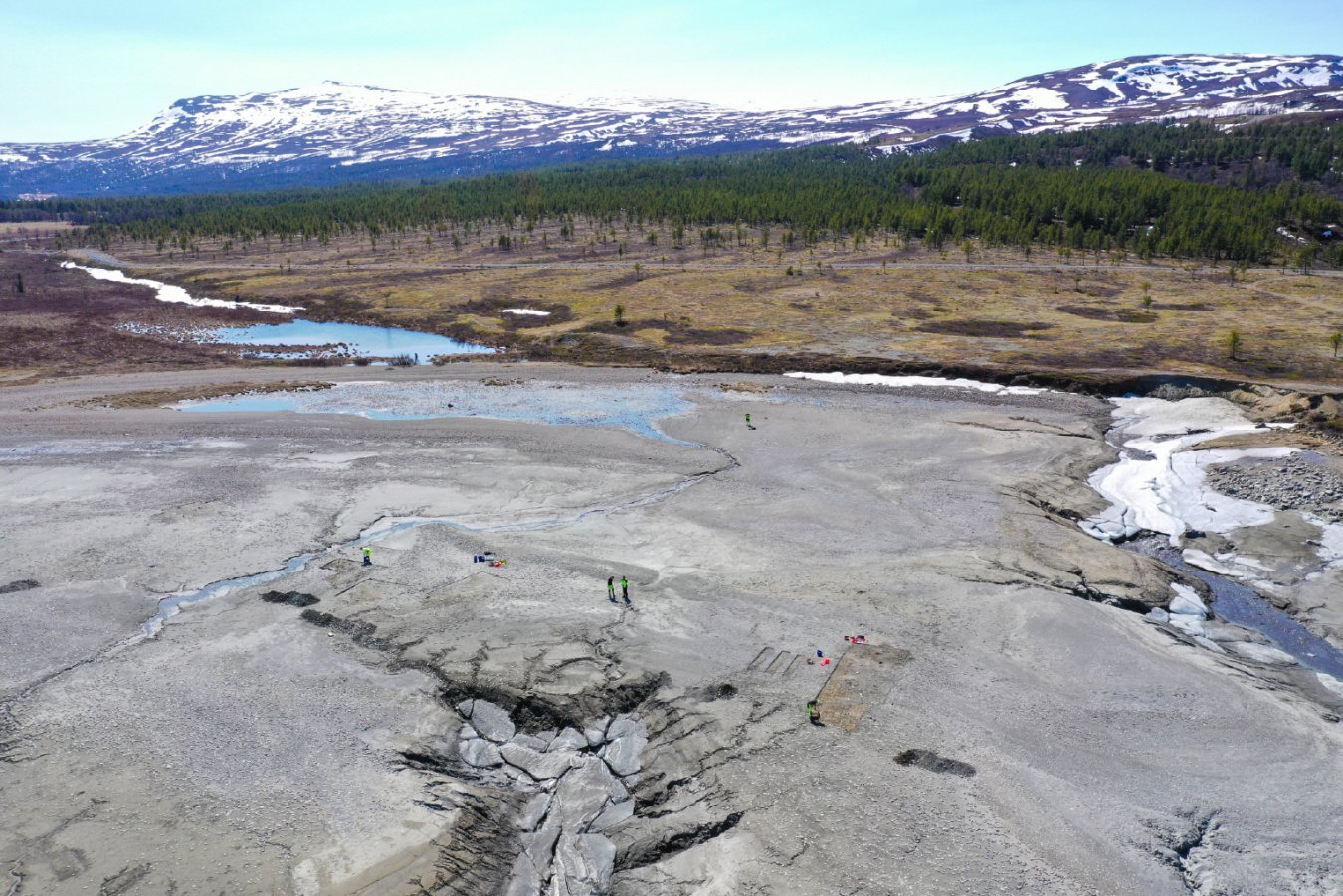 Aerial view of several small figures in high-visibility vests working on a cracked, debris-covered riverbed with snow-patched banks, set against a forested valley and snow-capped mountains.