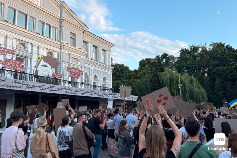 A crowd of people holding cardboard signs and a Ukrainian flag gathers outside a building displaying a portrait mural with Cyrillic text.