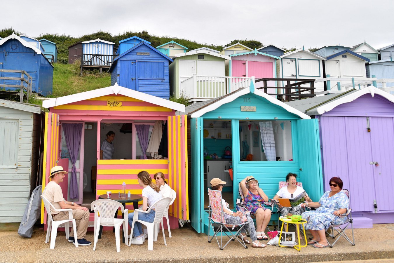 People sit in plastic chairs outside colorful wooden beach huts on a sandy area, with rows of blue, white, and pastel huts arrayed on a grassy hillside behind them.