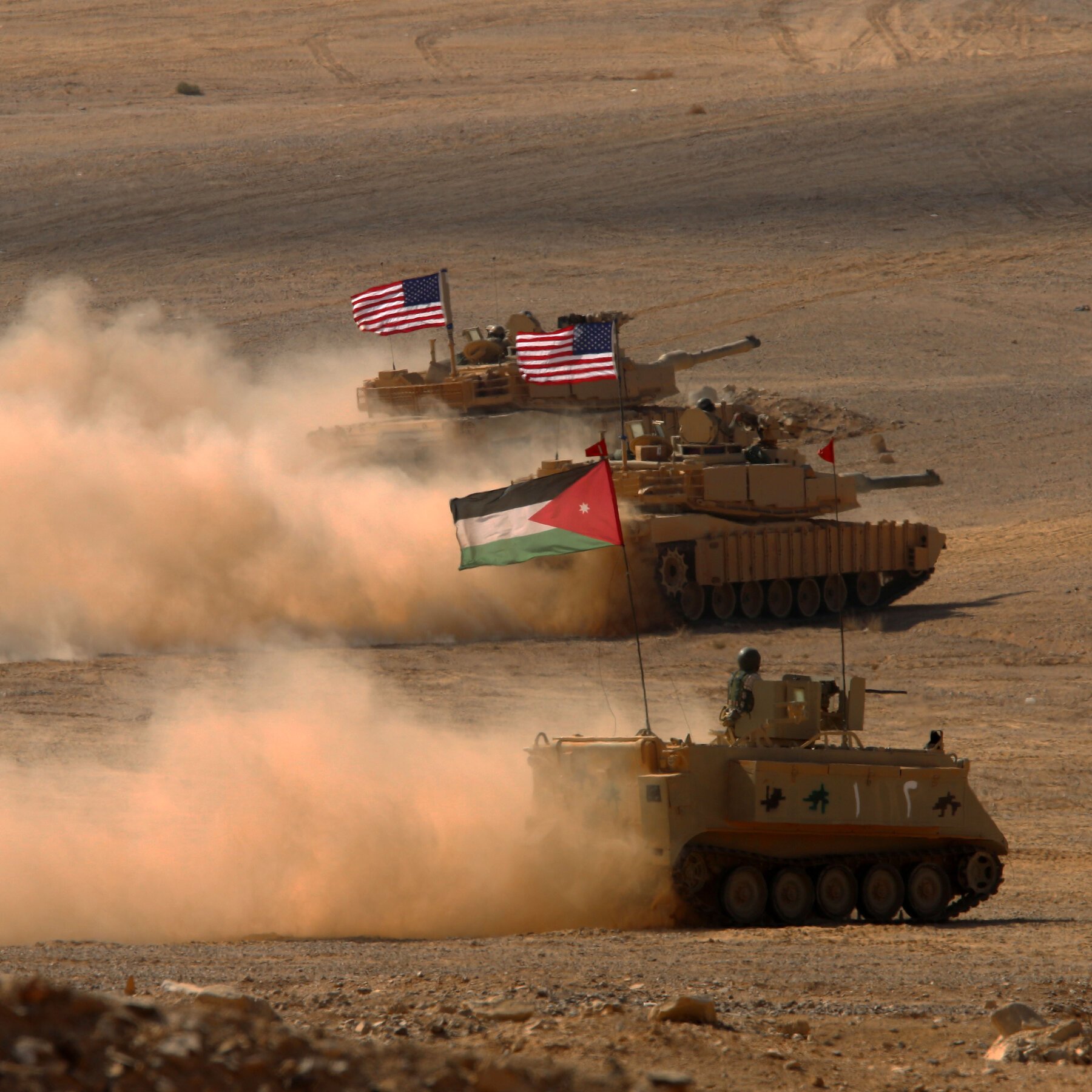 Three military vehicles with American and Jordanian flags drive across a dusty desert terrain, kicking up large clouds of sand.
