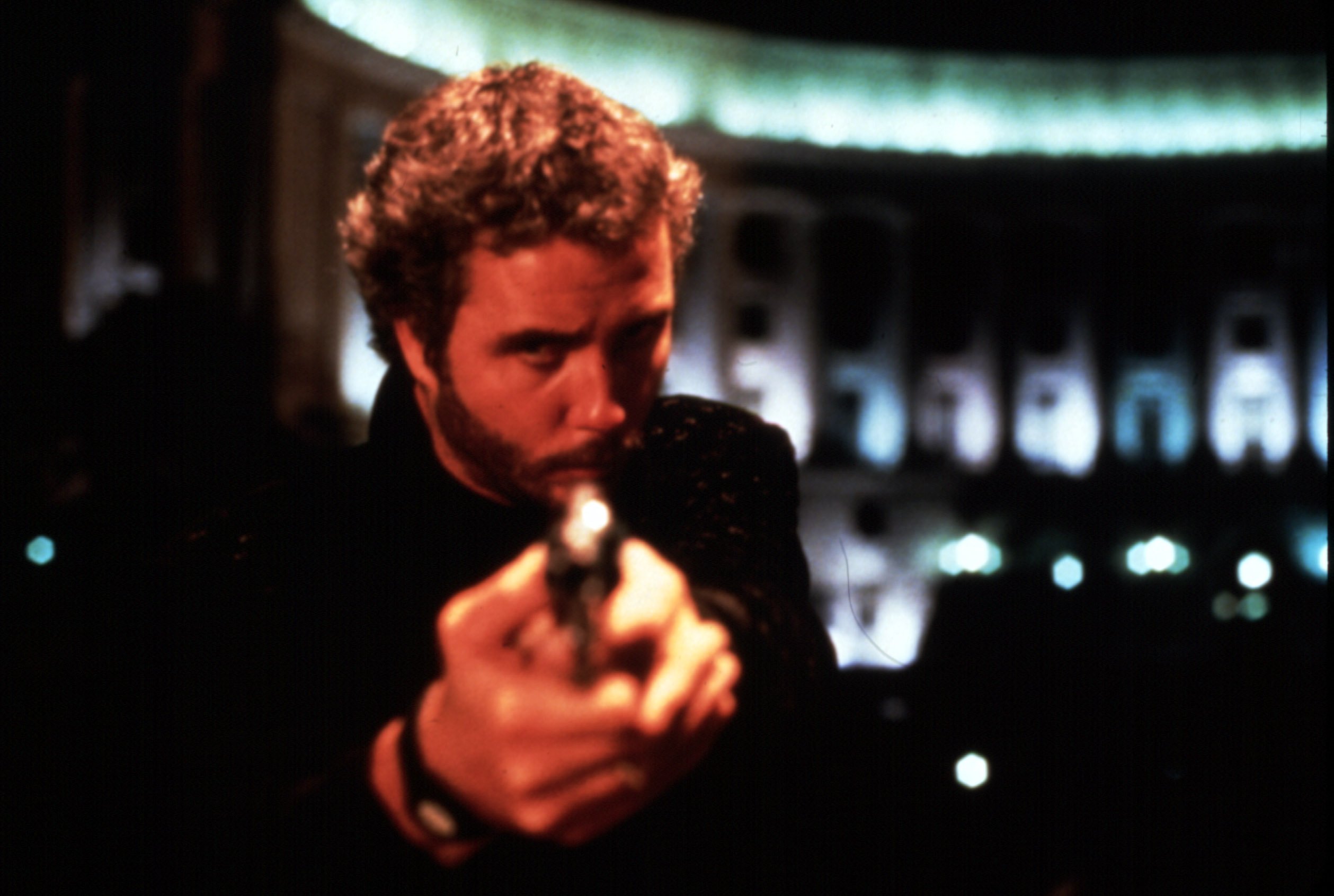 A curly-haired man in dark clothing aims a handgun forward in a dimly lit interior with arched, illuminated windows in the background.