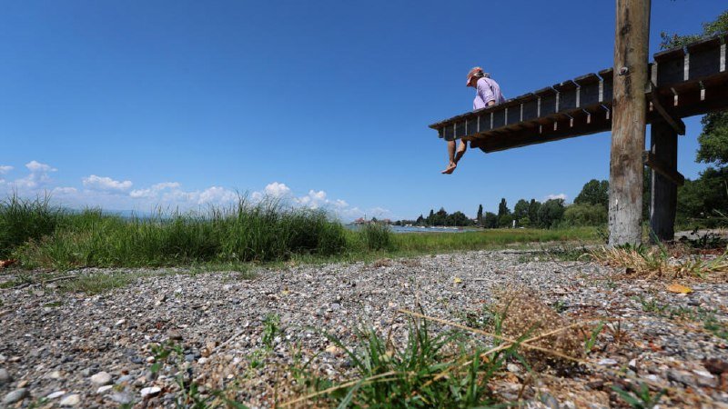 A person in a striped shirt and cap sits at the edge of an elevated wooden dock, dangling their legs above a pebbled shoreline with grass and water in the background.