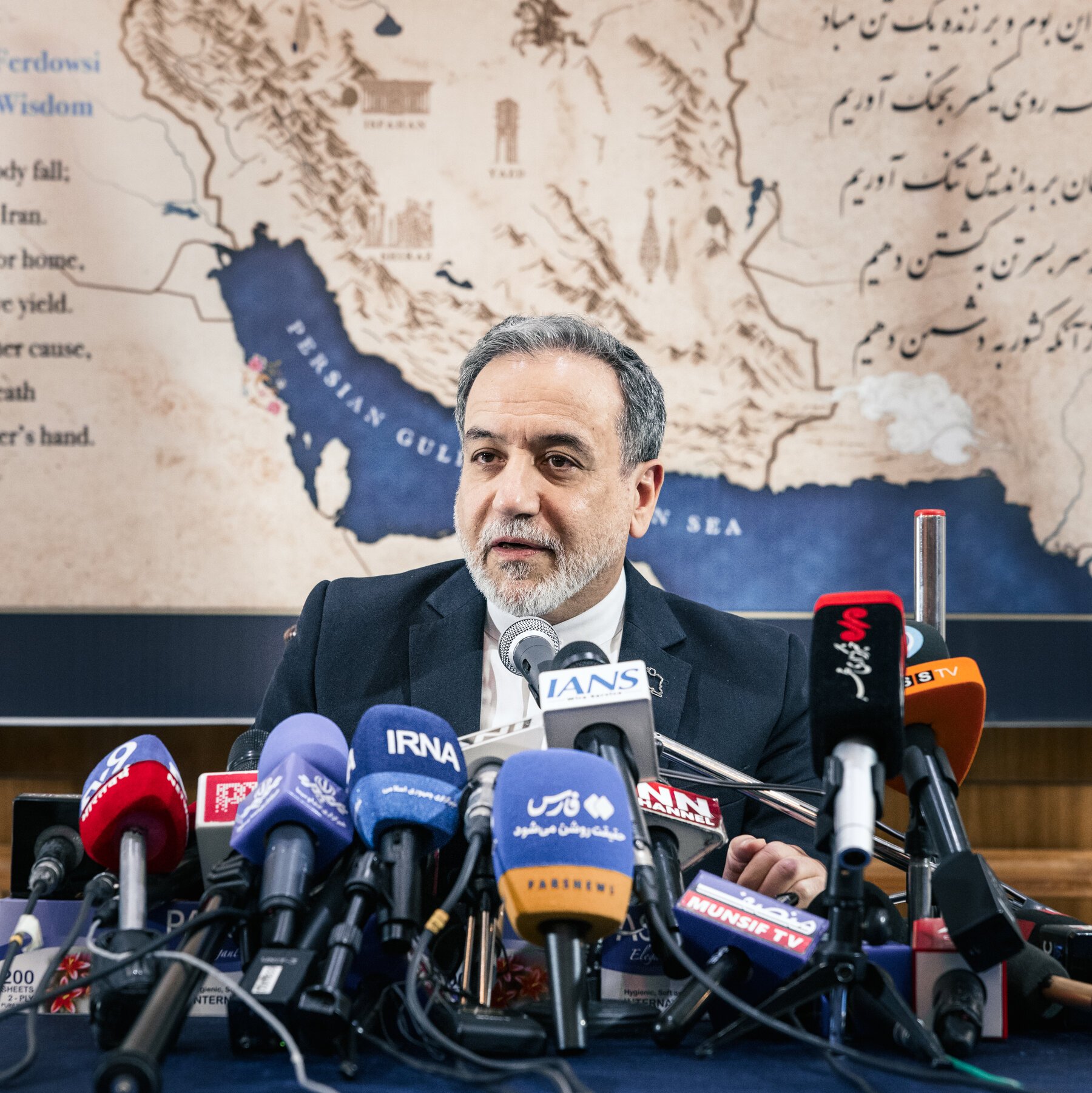 A gray-bearded man in a dark suit speaks behind a cluster of media microphones labeled IRNA, IANS, and others, before a backdrop displaying a map of the Persian Gulf region and Persian script.