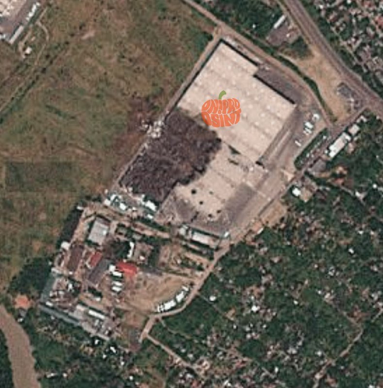 Satellite imagery shows a large industrial complex with a damaged warehouse structure, surrounded by cleared land, smaller buildings, and adjacent residential areas.