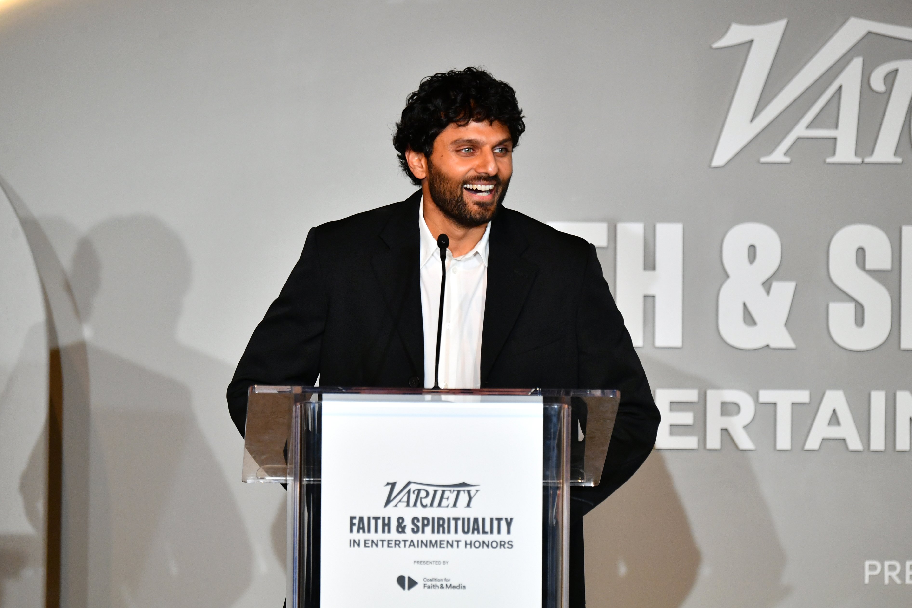 A smiling man in a black suit stands at a clear podium speaking into a microphone at the "Variety Faith & Spirituality in Entertainment Honors" event.