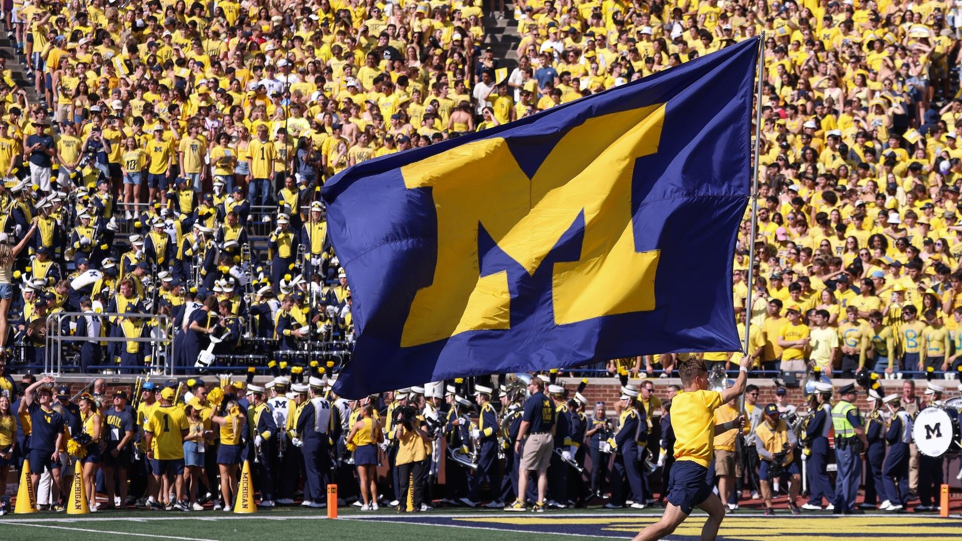 A man runs across a football field carrying a large blue and yellow "M" flag, with marching band members and a crowd wearing yellow shirts in the stands behind him.