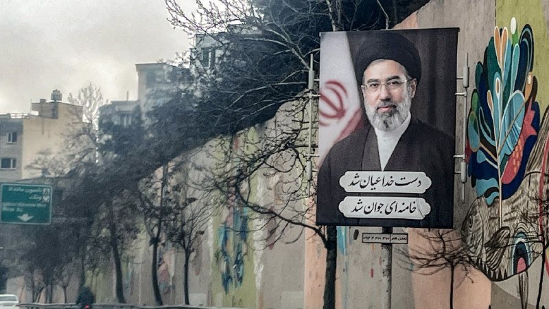 A large billboard displaying a portrait of a bearded man in clerical attire and a black turban is mounted on a wall, with Persian text and an Iranian flag visible.