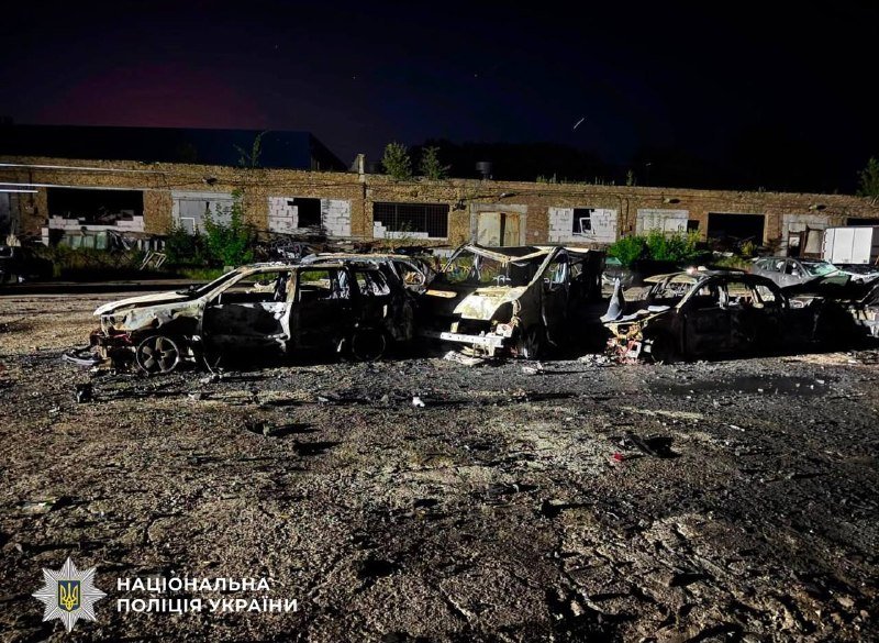 Multiple burnt and destroyed vehicles sit in a debris-strewn lot in front of a damaged brick building at night, with a "Національна поліція України" logo visible.
