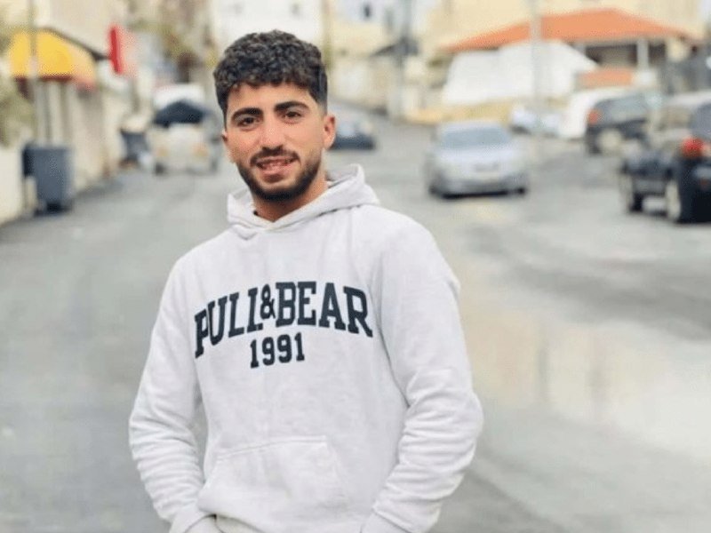 A young man with a beard stands smiling on a street, wearing a white "Pull&Bear 1991" hoodie, with parked cars and buildings visible behind him.