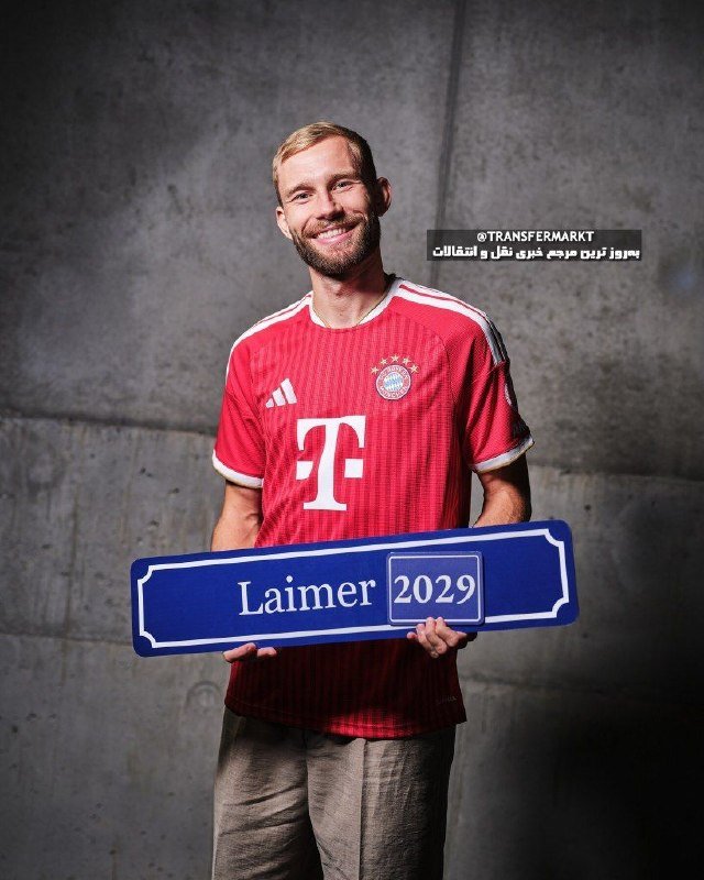 A smiling man in a red Bayern Munich jersey holds a blue sign reading "Laimer 2029" against a gray concrete backdrop.