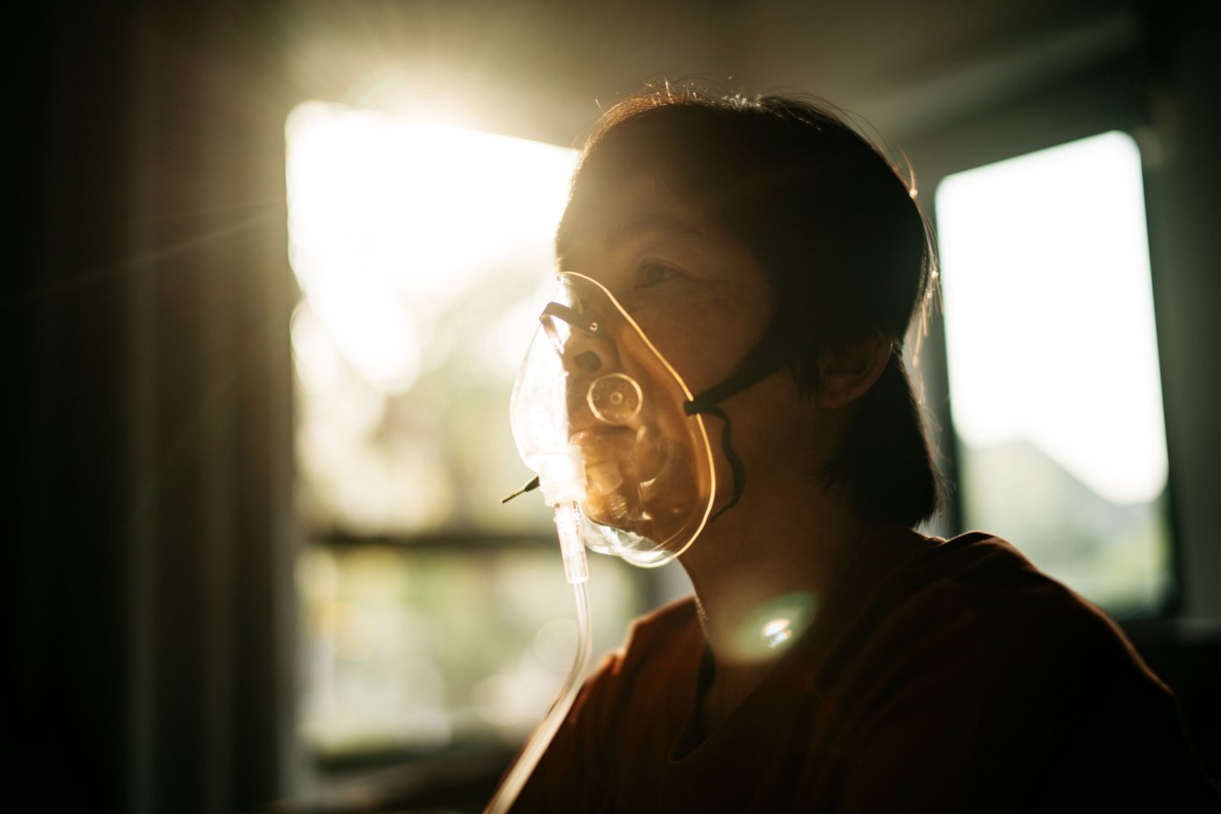 A person with short dark hair wearing a clear oxygen mask sits in profile, backlit by sunlight streaming through a window.