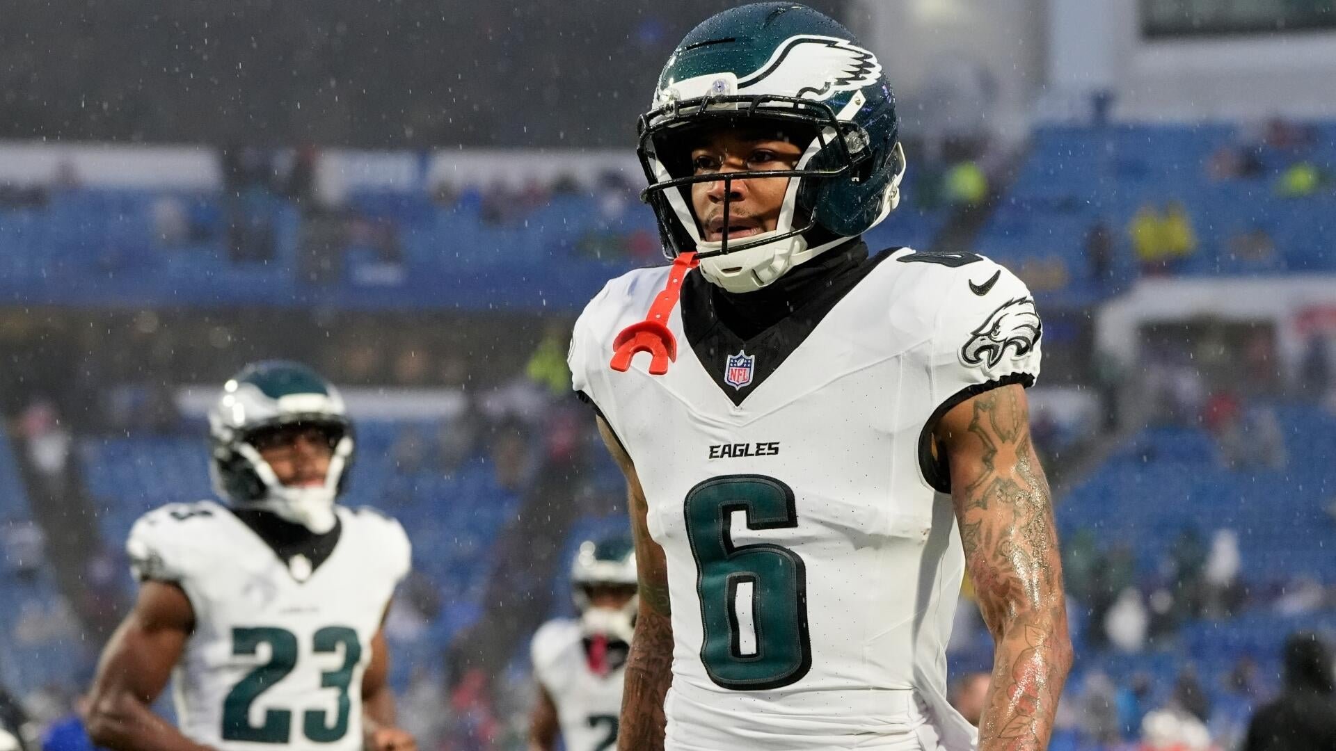 Philadelphia Eagles player number 6 walks on the field in a white jersey and helmet, with a red mouthguard hanging from his face mask, as rain falls visibly around him.