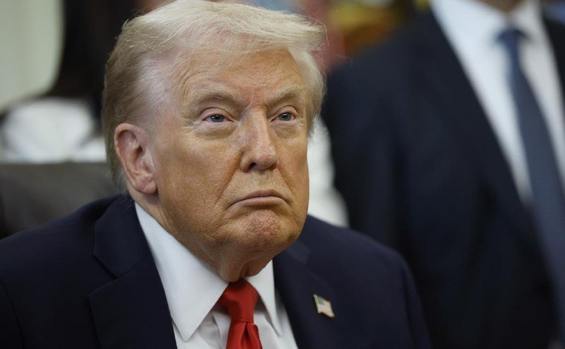 A blonde-haired man in a dark suit, white shirt, and red tie with an American flag pin sits with a serious expression, while another suited figure stands behind him.