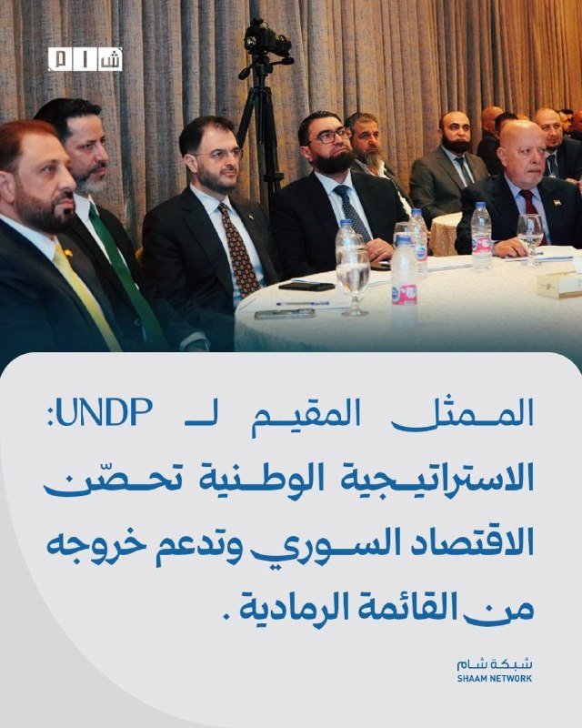 Men in suits seated at a conference table with water bottles, beneath Arabic text and a Shaam Network logo.