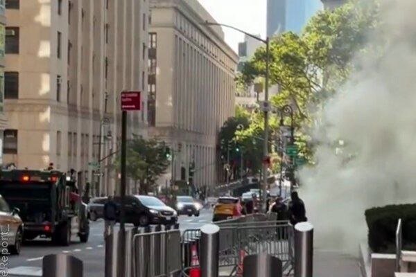A black armored vehicle and cars sit on a city street beside tall stone buildings, with plumes of white smoke rising along the sidewalk near pedestrians.