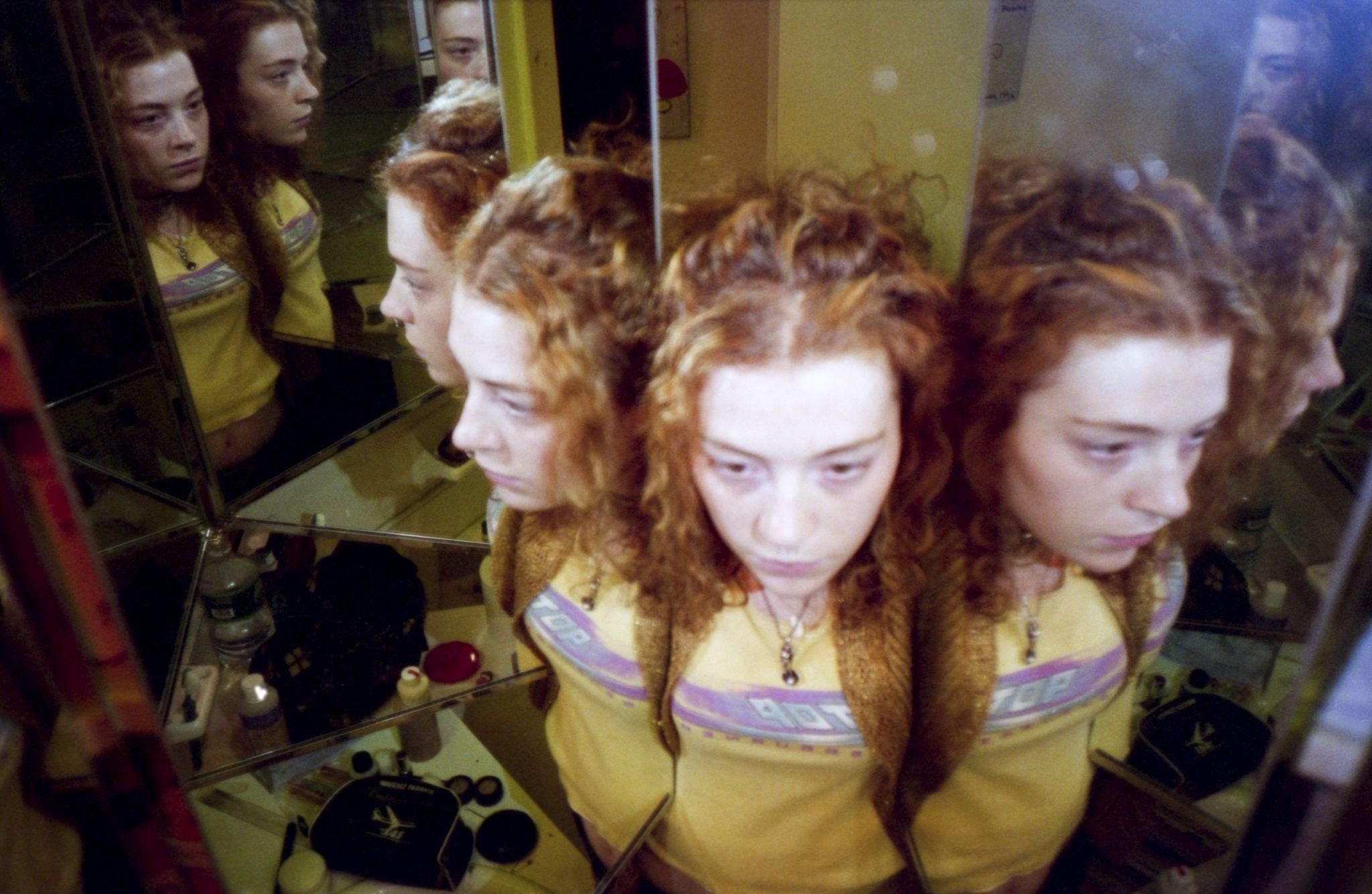 Multiple mirror reflections show a woman with curly red hair in a yellow top standing in front of a mirrored makeup counter.