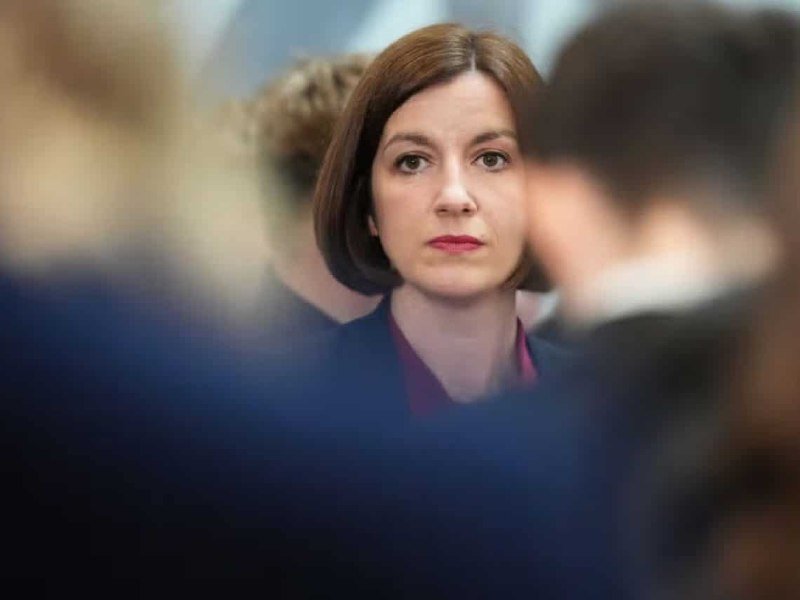 A woman with short brown hair and red lipstick stands among blurred figures in the foreground, looking forward with a serious expression.