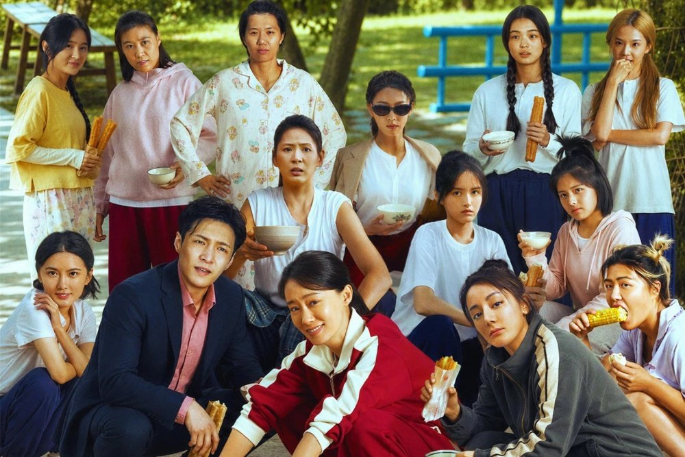 A group of thirteen people pose together outdoors, holding bowls and food items, with trees, a blue bench, and a picnic table visible in the background.