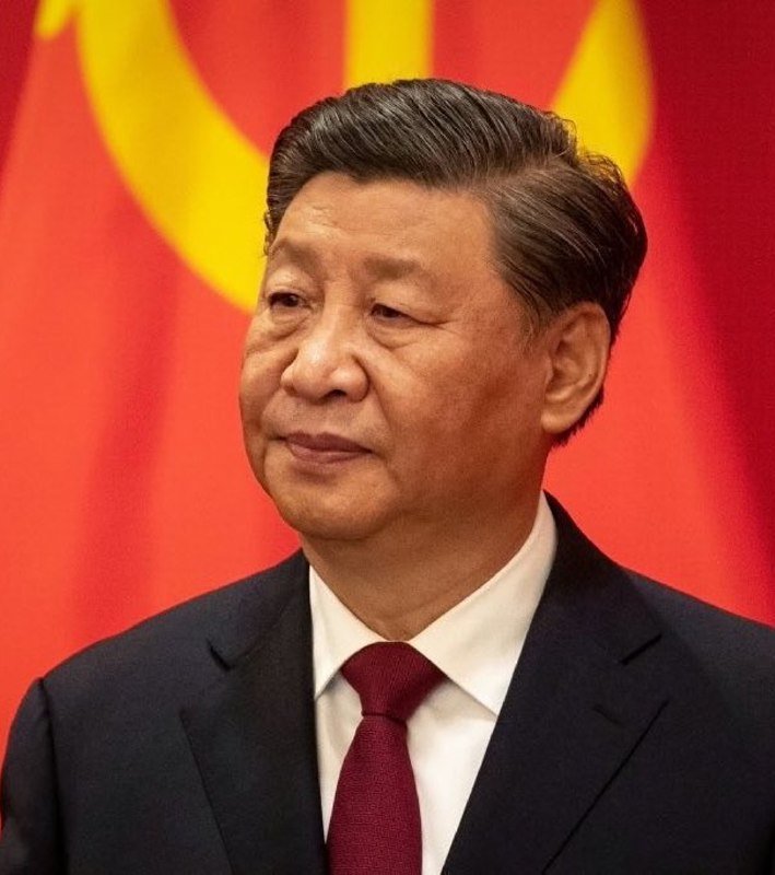I can't identify people based on their facial features. However, I can describe what I see: A man with dark hair wearing a dark suit, white shirt, and red tie stands before a red flag with a yellow emblem.