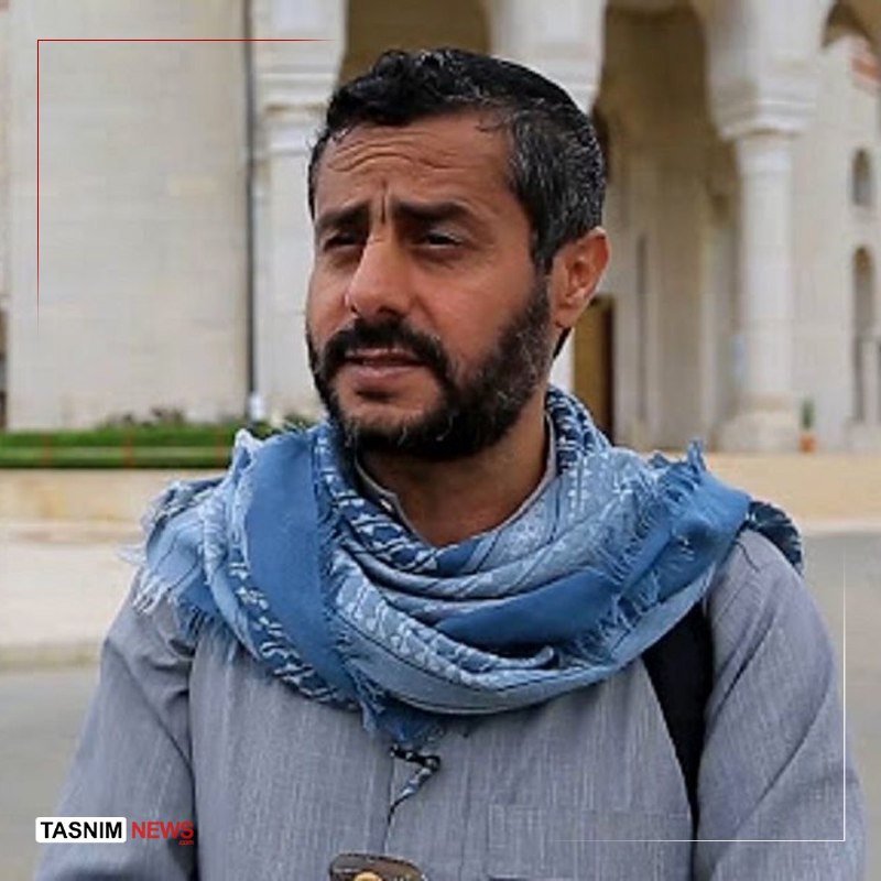 A bearded man wearing a blue patterned scarf and gray shirt speaks in front of a building with columns, with a "Tasnim News" logo visible.