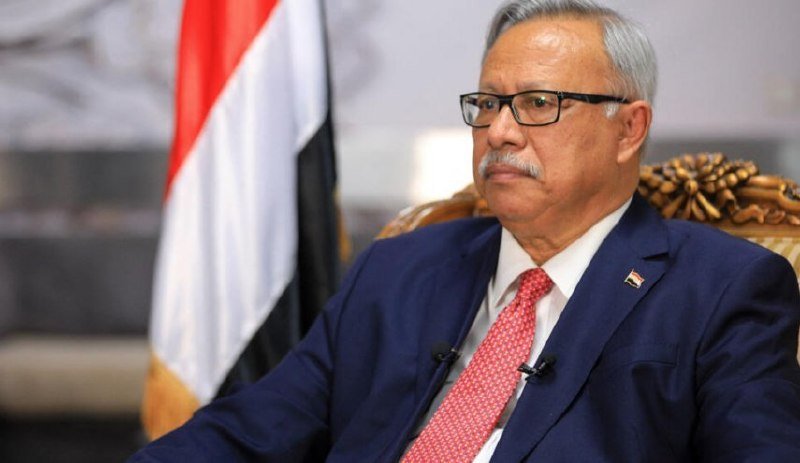 A gray-haired man in a blue suit, red patterned tie, and glasses sits in an ornate chair beside a red, white, and black flag.