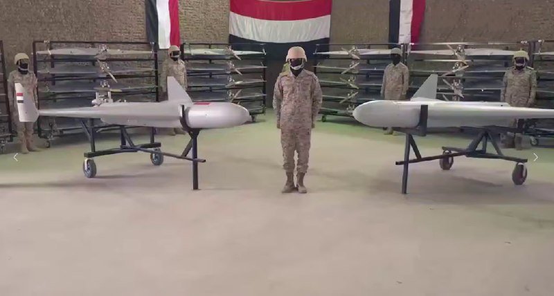Masked military personnel stand among displayed drones and missiles in an indoor facility, with red, white, and black flags visible in the background.