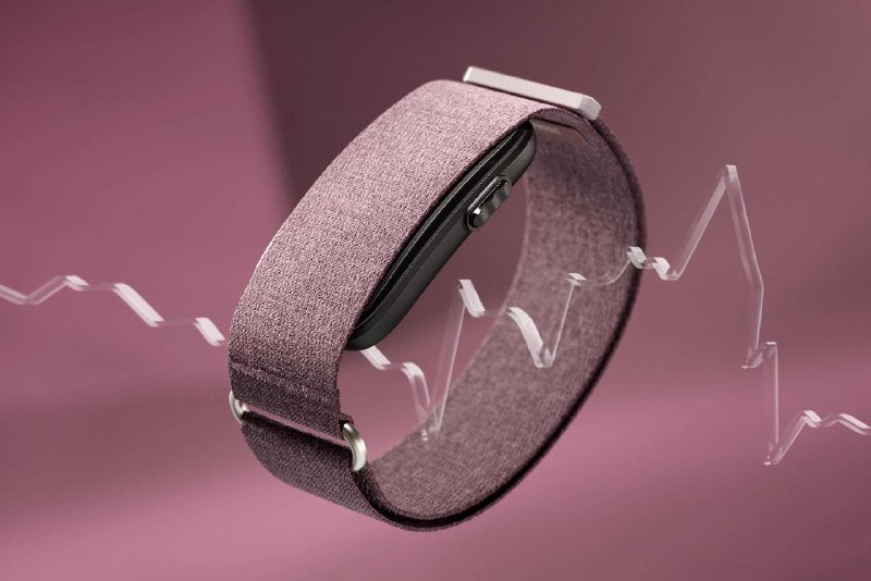 A fabric-banded smartwatch rests upright on a clear acrylic stand against a mauve background.
