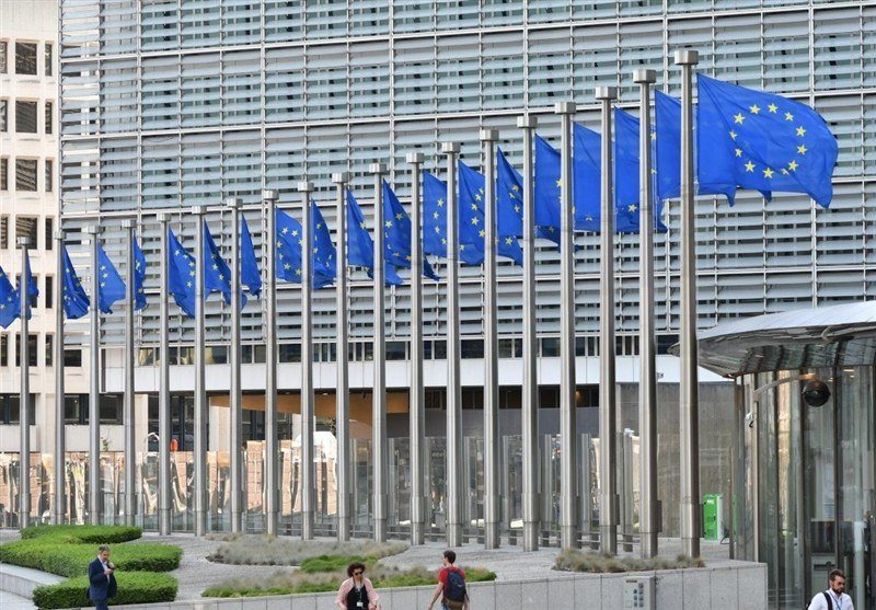A row of European Union flags flies on flagpoles outside a modern glass-and-steel office building, with pedestrians walking along the paved entrance area.