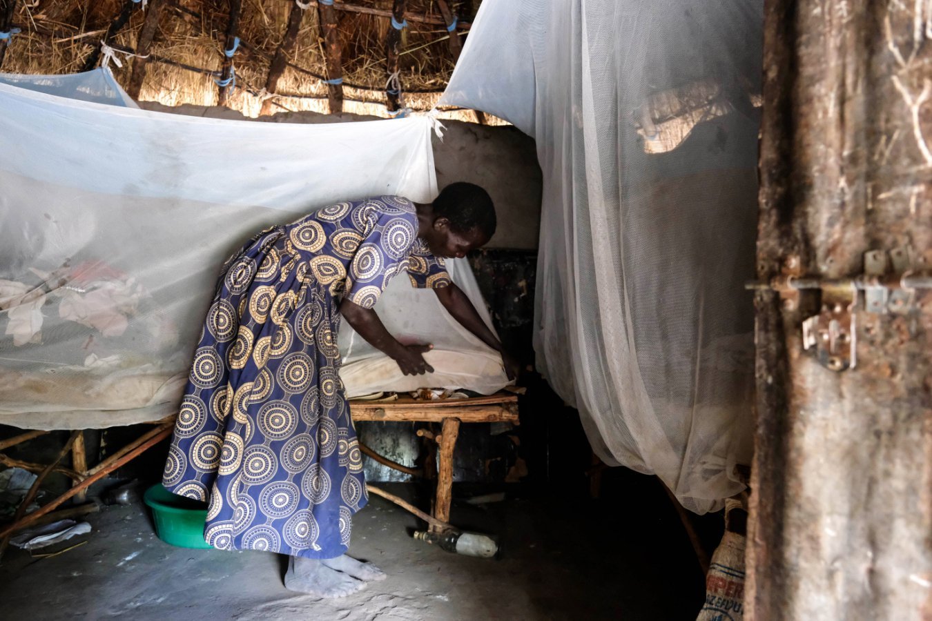 A woman in a patterned blue and yellow dress bends over to adjust a mosquito net draped around a wooden bed inside a thatch-roofed hut.