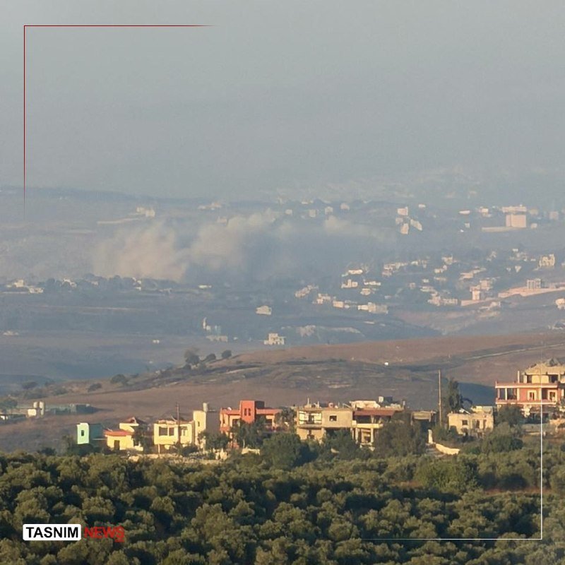A plume of smoke rises in the distance across a hazy valley of scattered buildings, viewed from an elevated vantage point above a cluster of houses and trees.