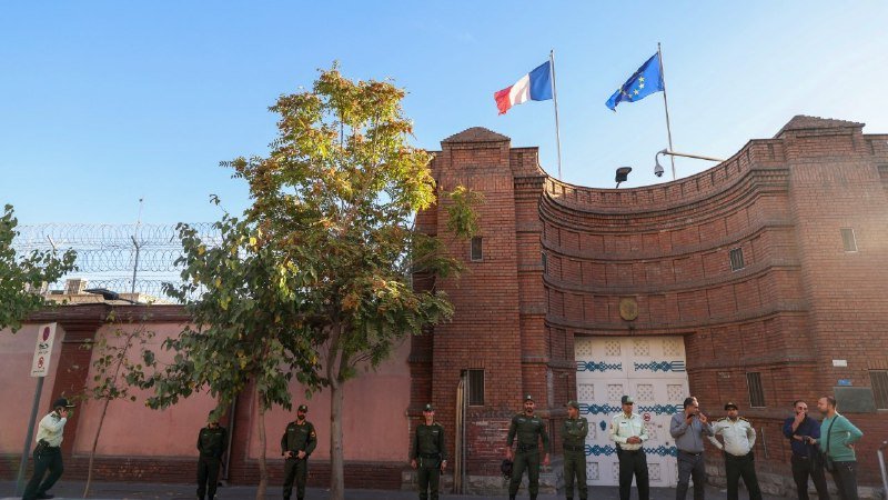 A large red-brick building flies French and European Union flags above its gated entrance, with uniformed officers standing watch along the perimeter wall beneath barbed wire.