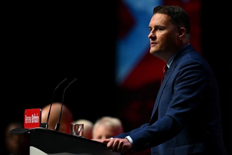 A man in a dark suit and red tie stands at a "Renew Britain" podium, speaking before an audience with a red, white, and blue flag in the background.