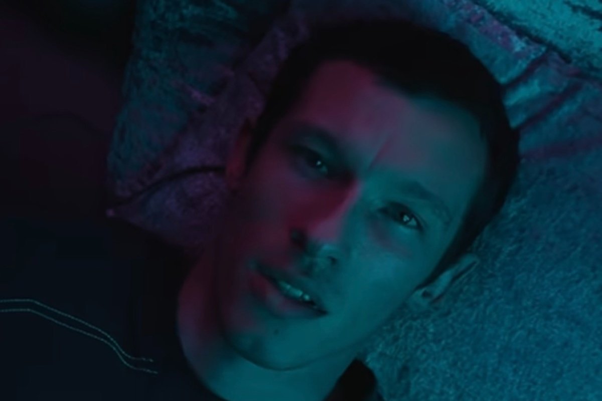 A close-up of a young man lying on a textured surface, illuminated by contrasting pink and cyan lighting.