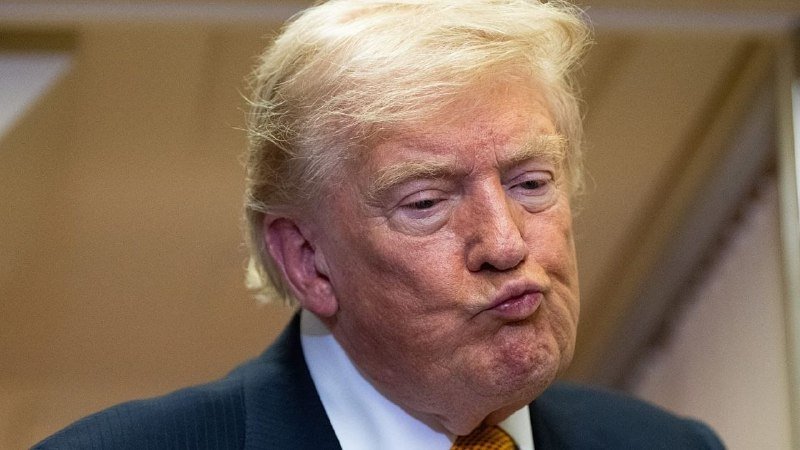 A man with blond hair wearing a dark suit, white shirt, and orange tie purses his lips with a serious expression against a beige background.