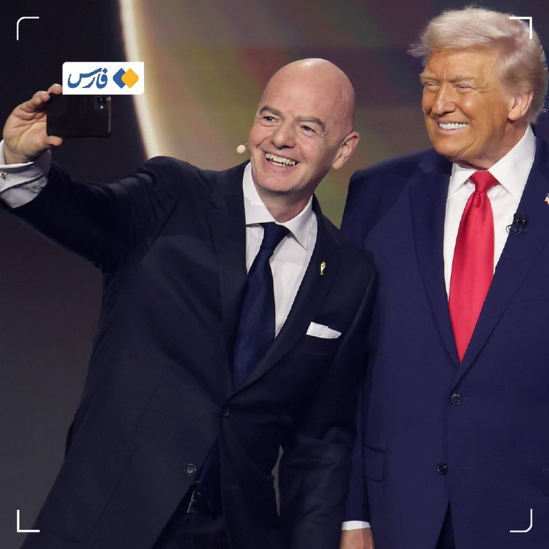 A bald man in a dark suit holds up a smartphone to take a selfie alongside a blonde-haired man in a blue suit and red tie, both smiling.