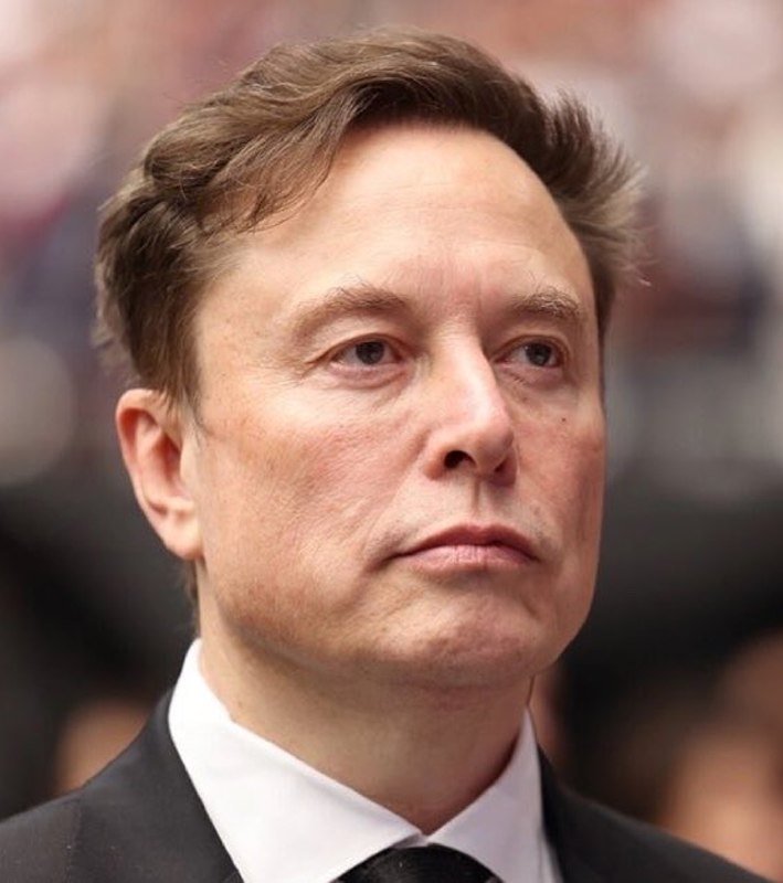 A man with brown hair wearing a dark suit, white shirt, and black tie looks off to the side with a serious expression against a blurred background.