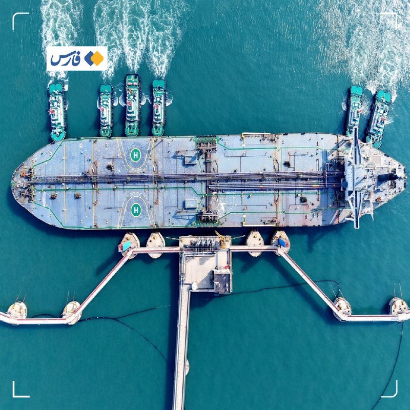 Aerial view of a large tanker ship docked at a marine terminal, with smaller tugboats maneuvering alongside in turquoise waters.