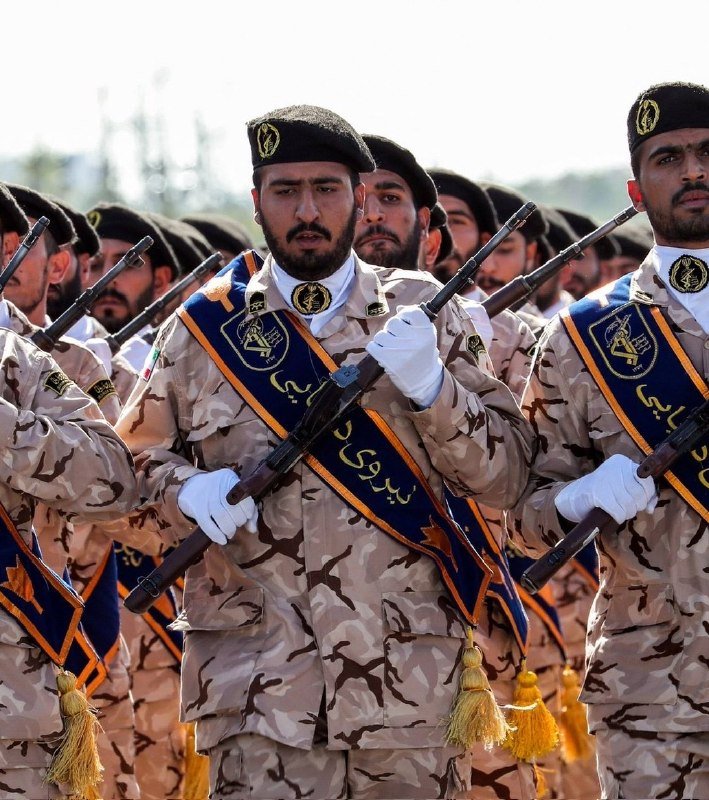 Soldiers in desert camouflage uniforms, black berets, and white gloves march in formation while holding rifles, wearing blue and yellow ceremonial sashes with tassels.