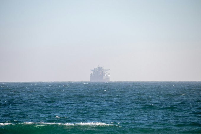 A large ship sits on the horizon across choppy blue ocean waters under a hazy, pale sky.