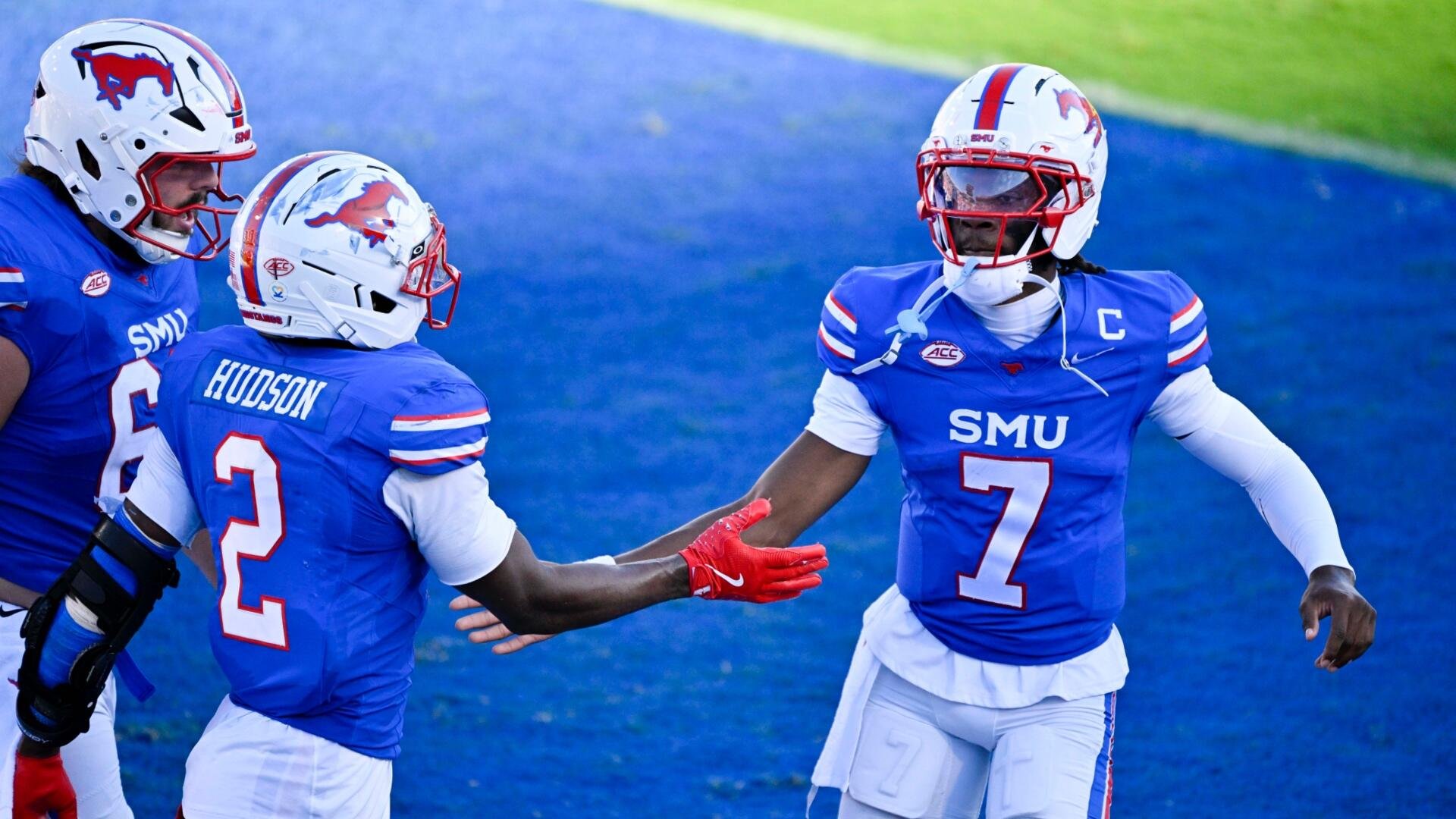 Three SMU football players in blue jerseys and red-white helmets celebrate on a blue field, with player #7 extending a red-gloved hand toward player #2 "Hudson."