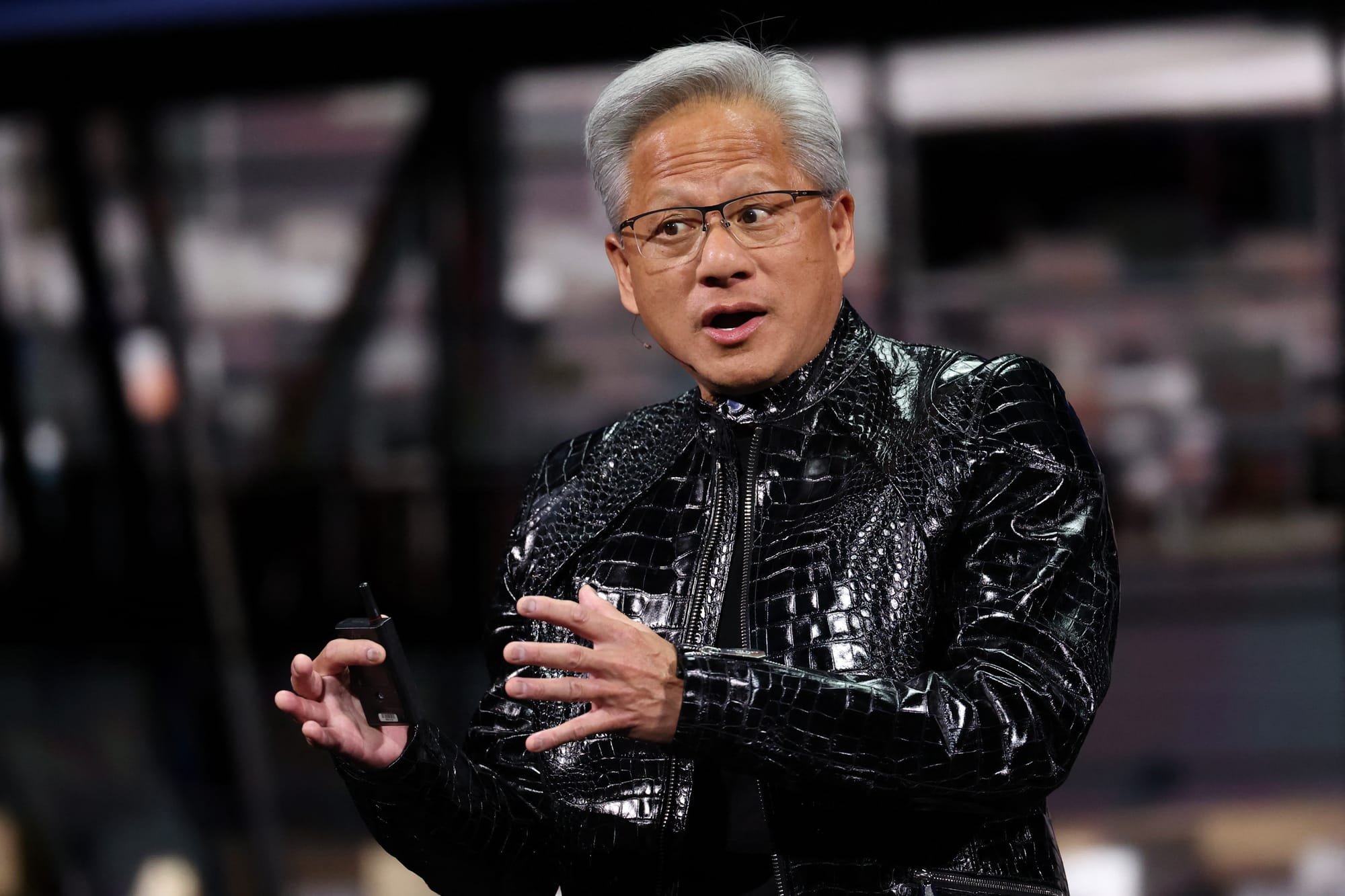 A man with silver hair and glasses, wearing a black textured crocodile-pattern jacket, gestures with both hands while holding a small device on a stage.