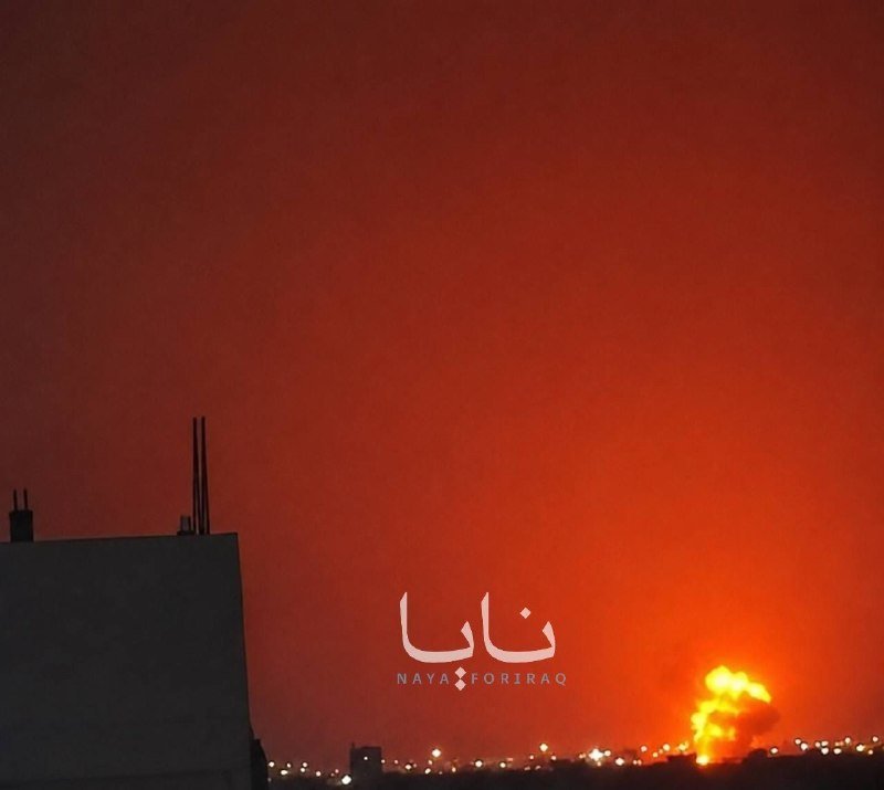 A bright orange explosion illuminates the night sky above a distant city skyline, with a building silhouette in the foreground and "نايا NAYA FORIRAQ" text overlaid.