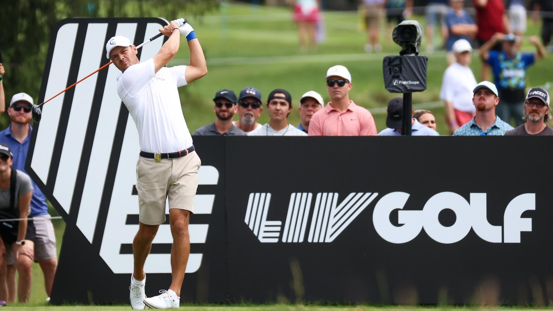 A golfer in a white shirt and khaki shorts completes his swing at a tee box, with a "LIV Golf" billboard and a crowd of spectators watching from behind.