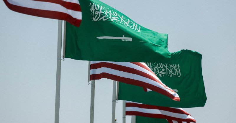 Multiple American and Saudi Arabian flags wave against a clear sky, mounted on flagpoles in an alternating arrangement.